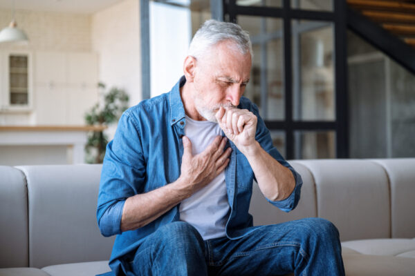 Senior man sitting on sofa at home and holding hand on chest. Male having asthma attack, difficulties with breathing, feeling severe pain or dyspnea