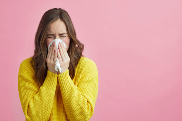 Woman in a yellow sweater is blowing her nose in front of a pink backdrop