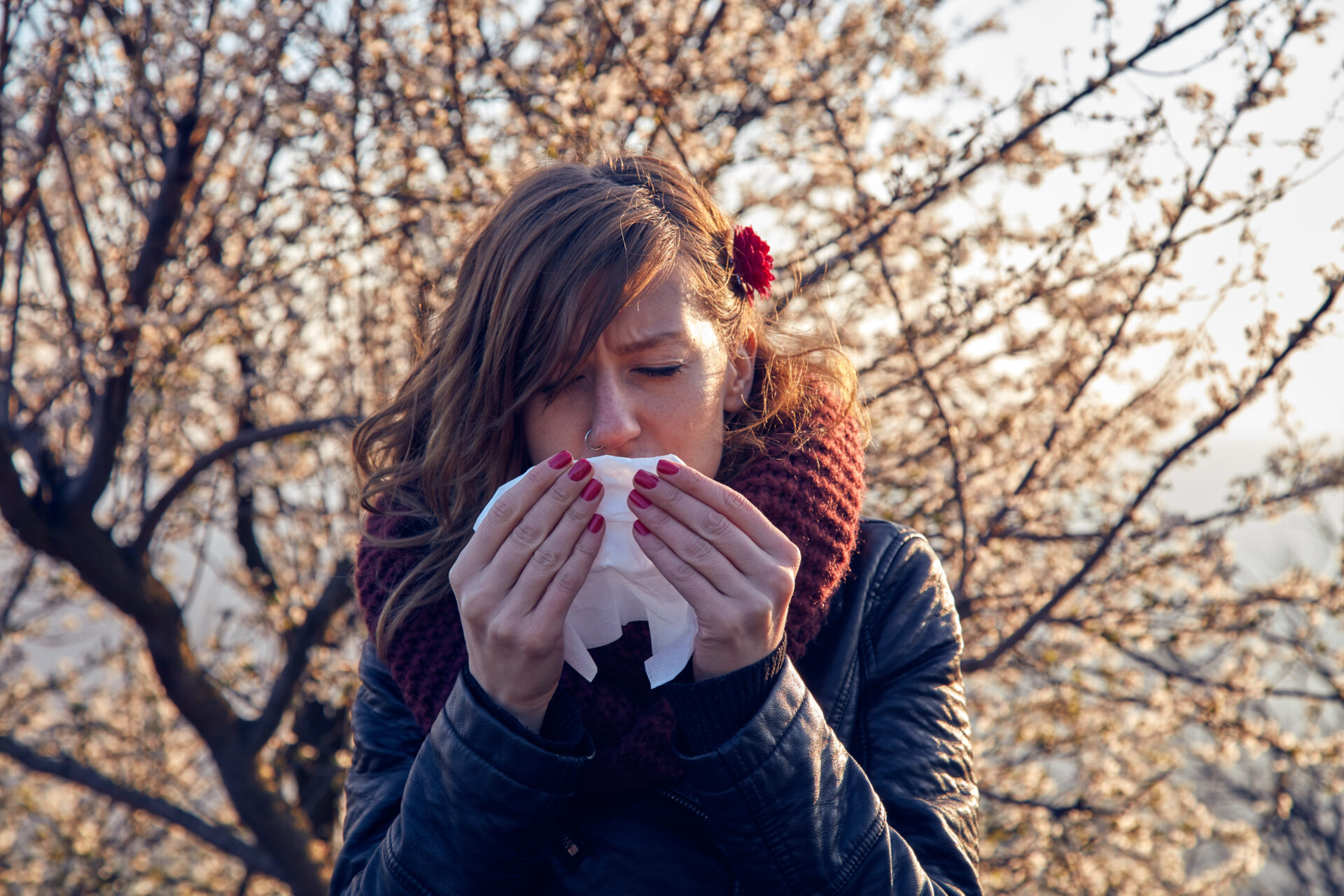Woman outside in a coat is sneezing into a tissue. A tree blossoms behind her.