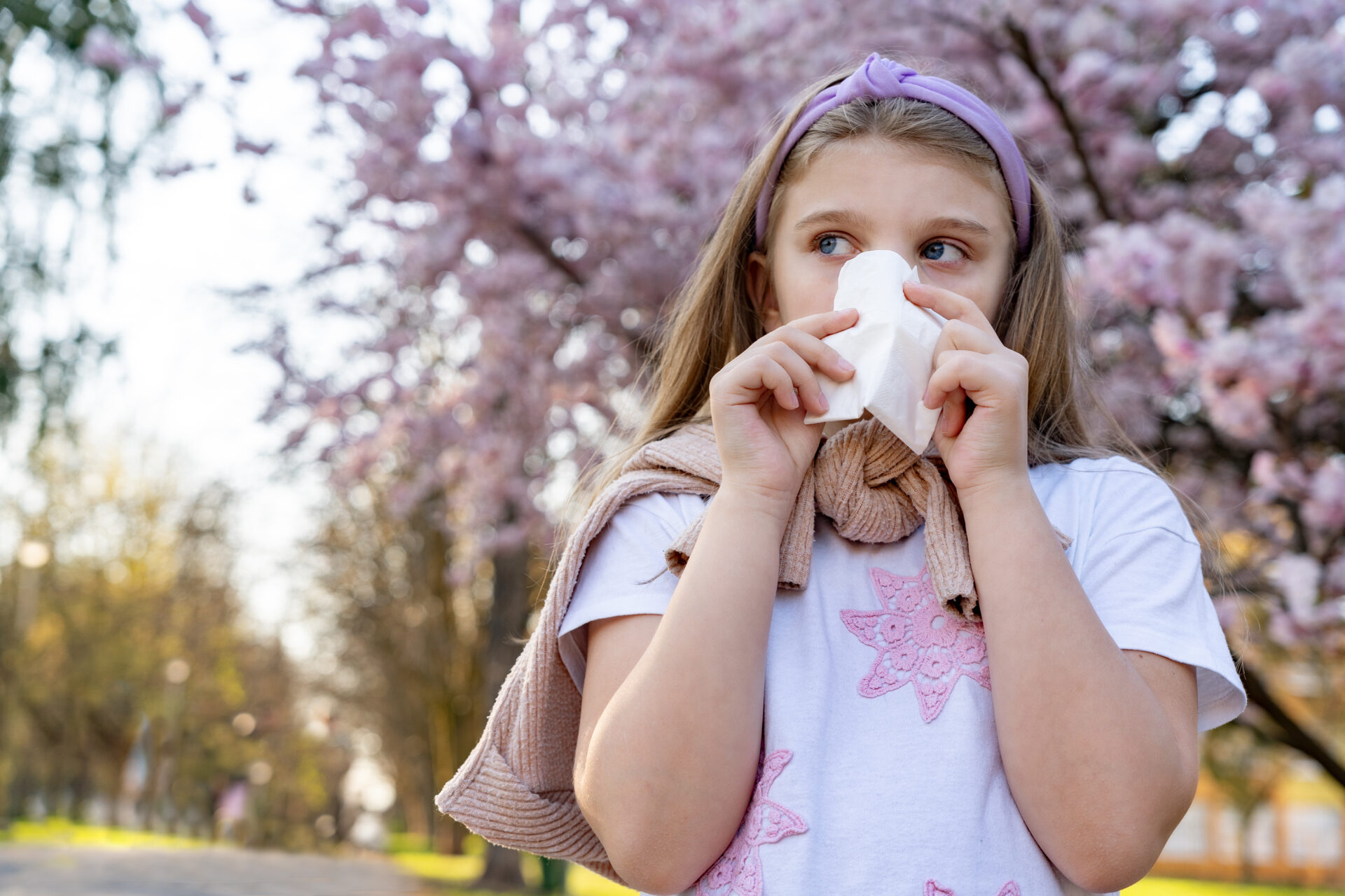 A young girl is walking down the street blowing her nose. A tree with pink flower blossoms is behind her.