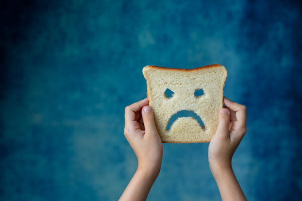 A child holds up a slice of bread with a frowny face carved into it--indicates food intolerance or food allergy.